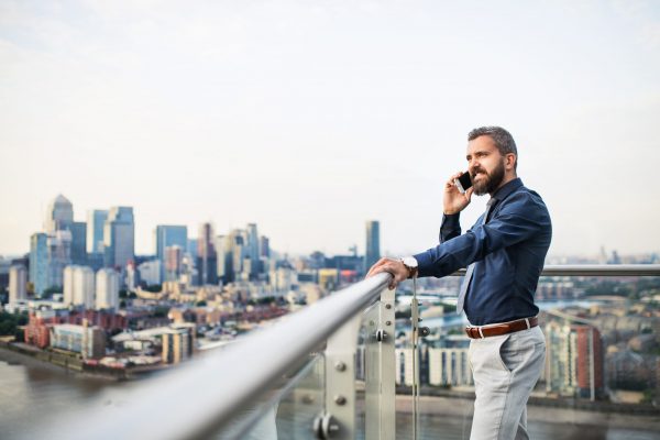 A portrait of businessman with smartphone standing against London rooftop view panorama, making a phone call. Copy space.